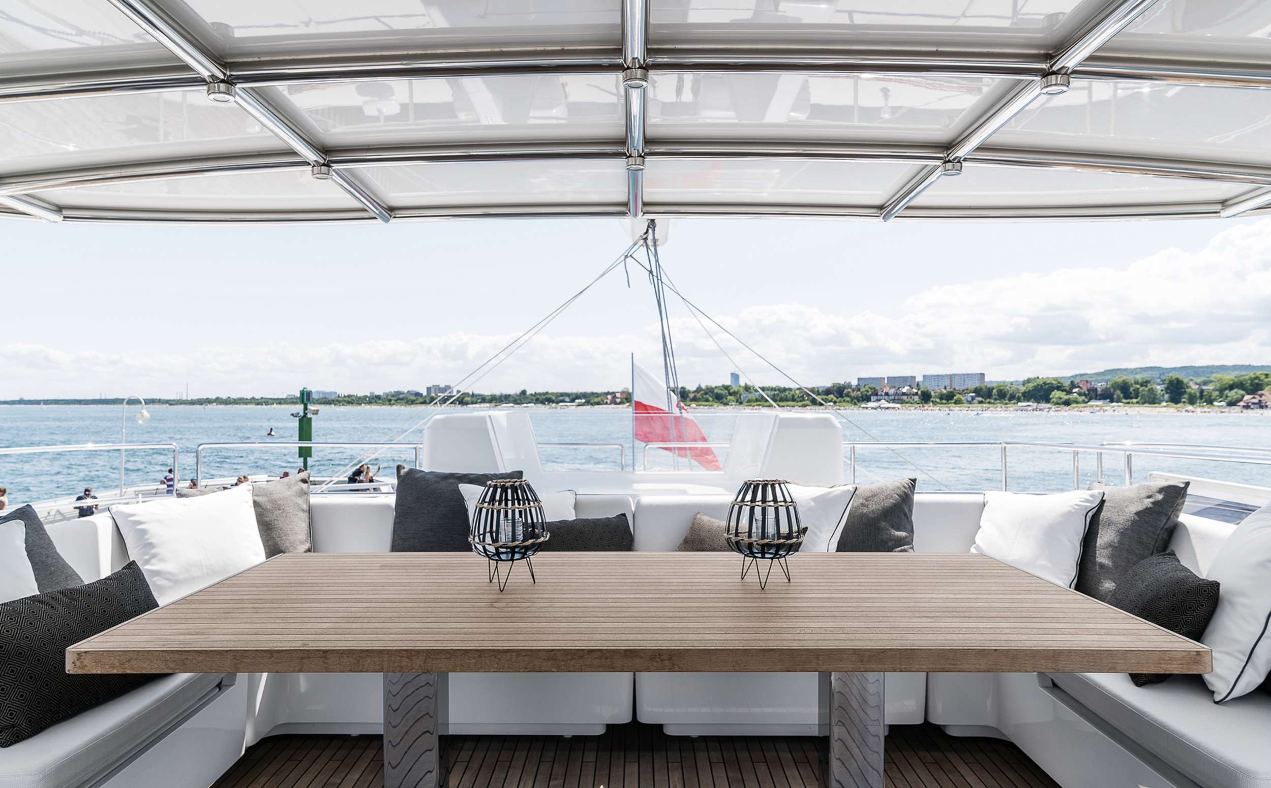 ABOVE & BEYOND A modern yacht deck with a wooden table, decorative lanterns, and cushioned seating, overlooking calm water and a distant shoreline under a partly cloudy sky. A flag is visible at the back of the yacht.