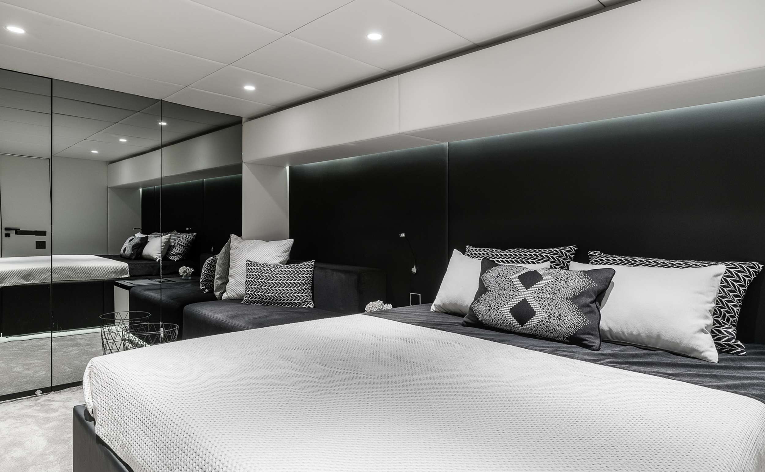 ABOVE & BEYOND Modern bedroom with a minimalist design, featuring a large bed with patterned pillows, a black accent wall, mirrored closet doors, a gray sofa, and soft overhead lighting.