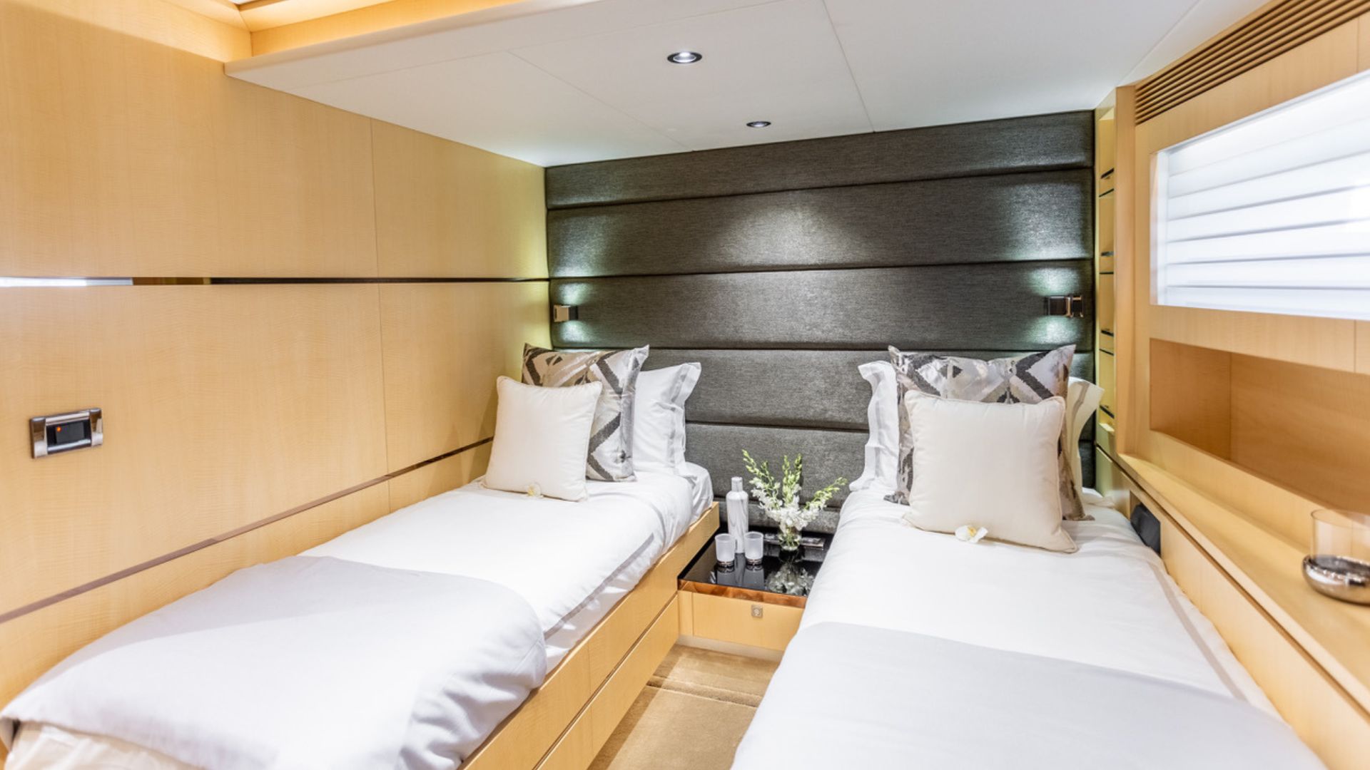 A modern, well-lit bedroom with two single beds separated by a small nightstand. Light wooden walls, white bedding, decorative pillows, and a dark accent wall create a cozy and elegant atmosphere.