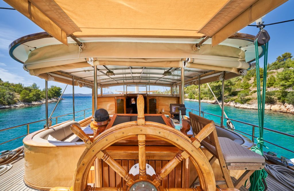 LIBRA A wooden ship’s helm with a shaded seating area is seen from the back, cruising through clear turquoise waters with lush green trees on the shoreline under a sunny sky.