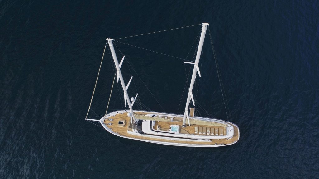Aerial view of a large white sailing yacht with wooden deck floating on dark blue water, featuring two masts, sun loungers, and a small pool or hot tub on deck.