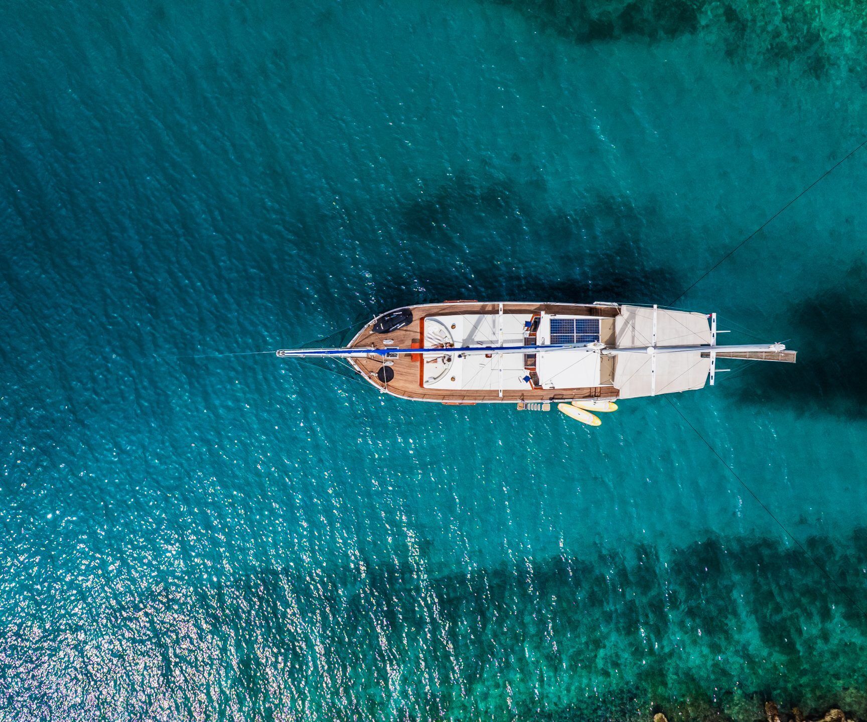 Aerial view of a sailboat anchored in clear, turquoise water near a shoreline. The boat casts a shadow on the sea, and sunlight sparkles on the waters surface.