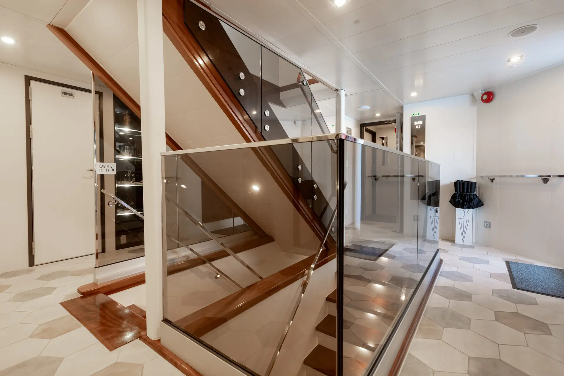 Modern indoor staircase with wooden steps and glass railings, located in a bright hallway with hexagonal tile flooring, white walls, and recessed ceiling lights. A closed white door and coat racks are visible.