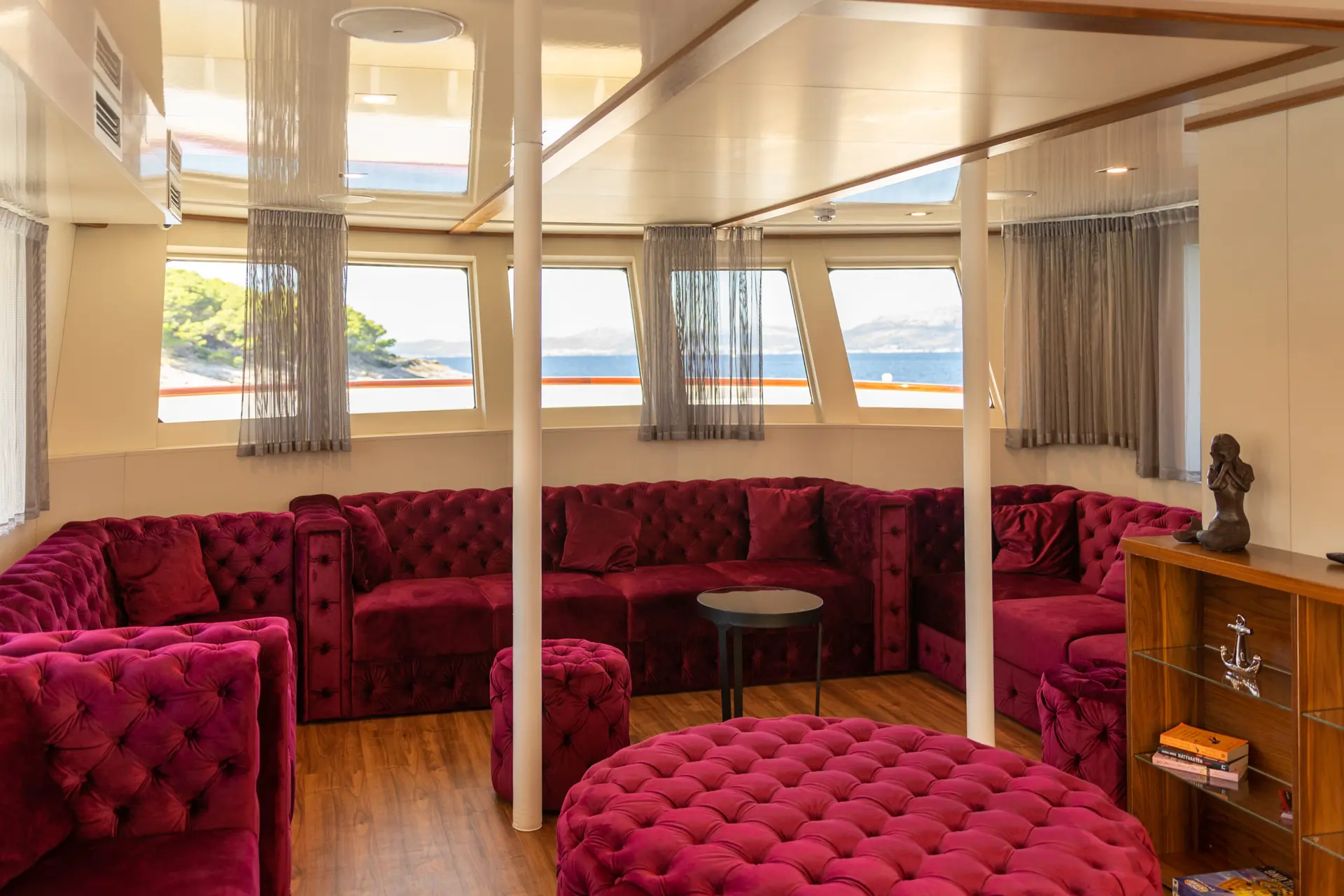 A cozy yacht lounge with plush, deep-red velvet sofas and round ottomans, wood flooring, windows with sea views, and natural light filling the space. A small bookshelf and a sculpture decorate one corner.