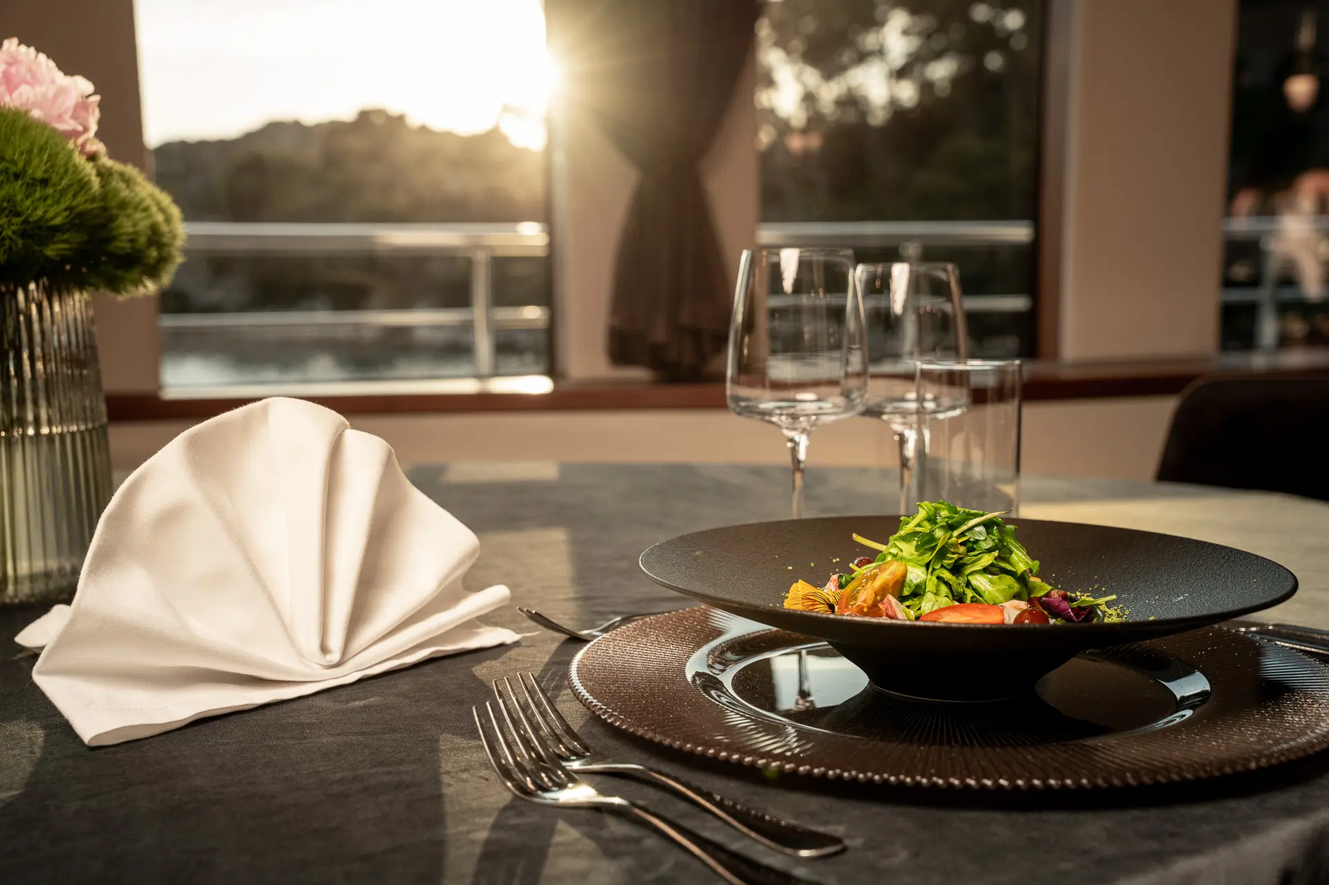 A sunlit dining table set with a folded white napkin, cutlery, empty wine glasses, and a black plate with a colorful salad. A flower vase and large windows with an outdoor view are in the background.