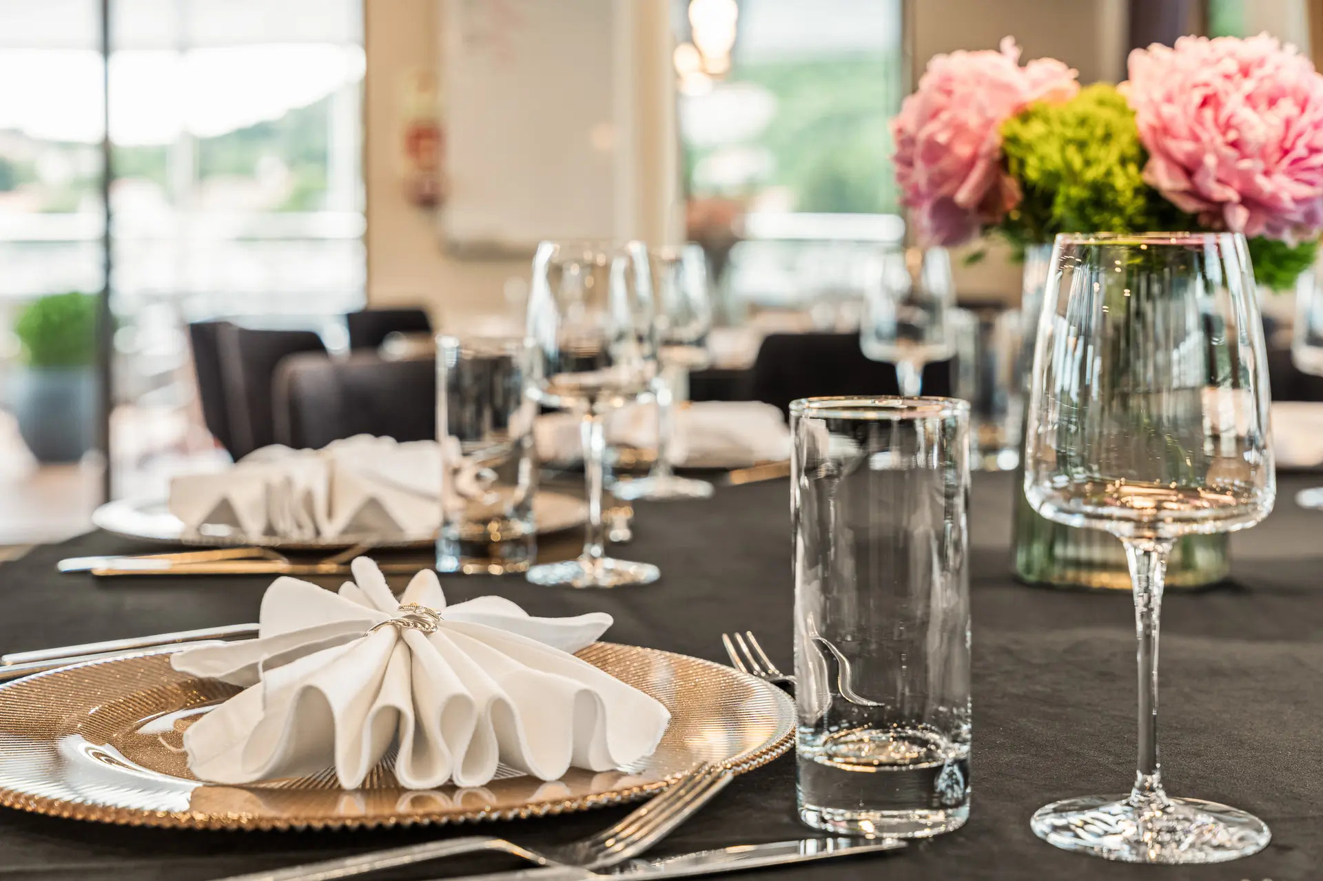 A close-up of an elegant dining table set with folded white napkins on gold plates, glassware, silver cutlery, and a floral centerpiece with pink and green flowers. The background shows a bright, blurred dining area.
