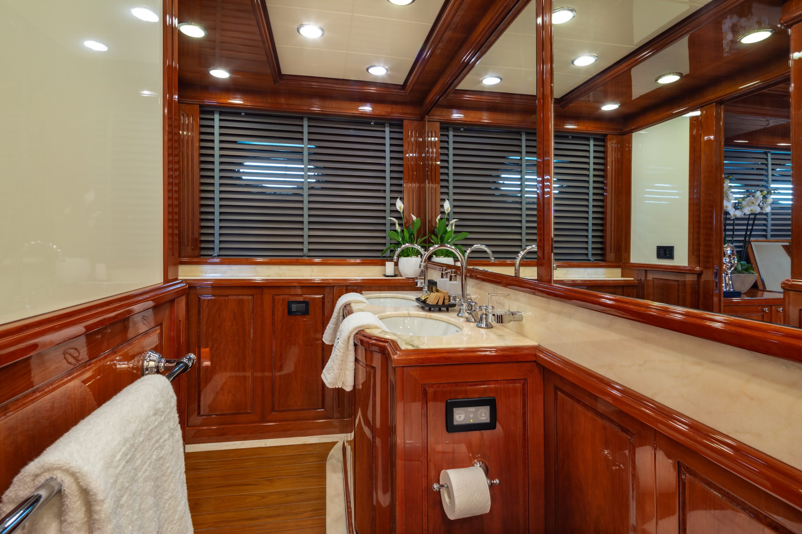 DIVINE Luxurious bathroom with polished wood cabinets, cream marble countertop, double sinks, large mirrors, recessed ceiling lights, a towel rack with a white towel, blinds on the window, and potted plants near the sinks.