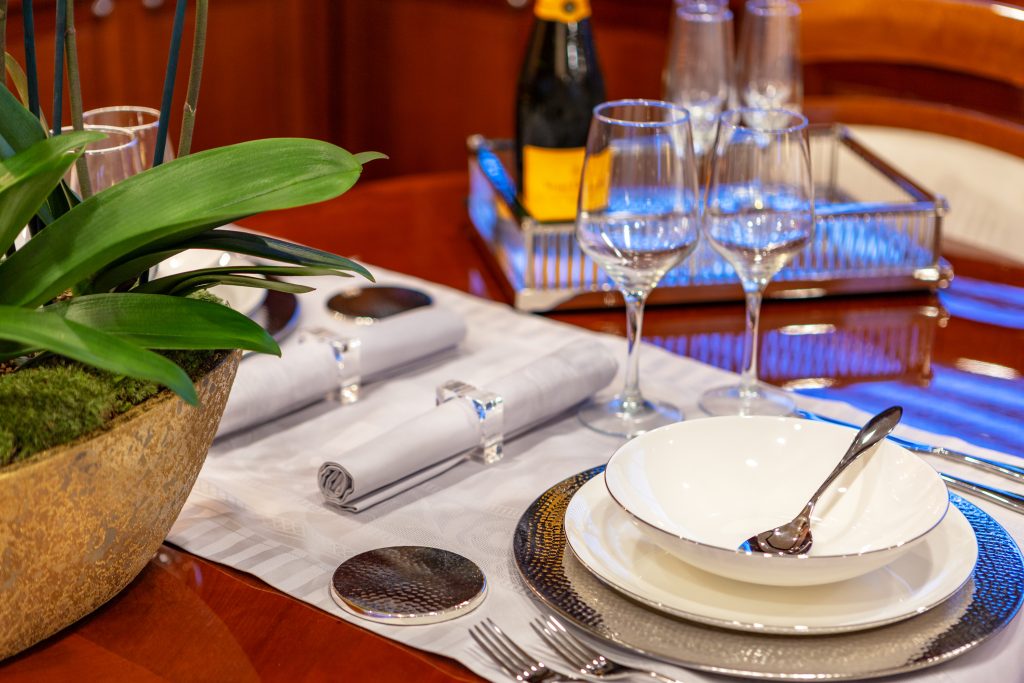 DIVINE A formal table setting with a white bowl, plate, spoon, fork, and knife on a silver placemat. Two rolled napkins with silver rings, wine glasses, a champagne bottle, and a green plant are also on the polished wooden table.