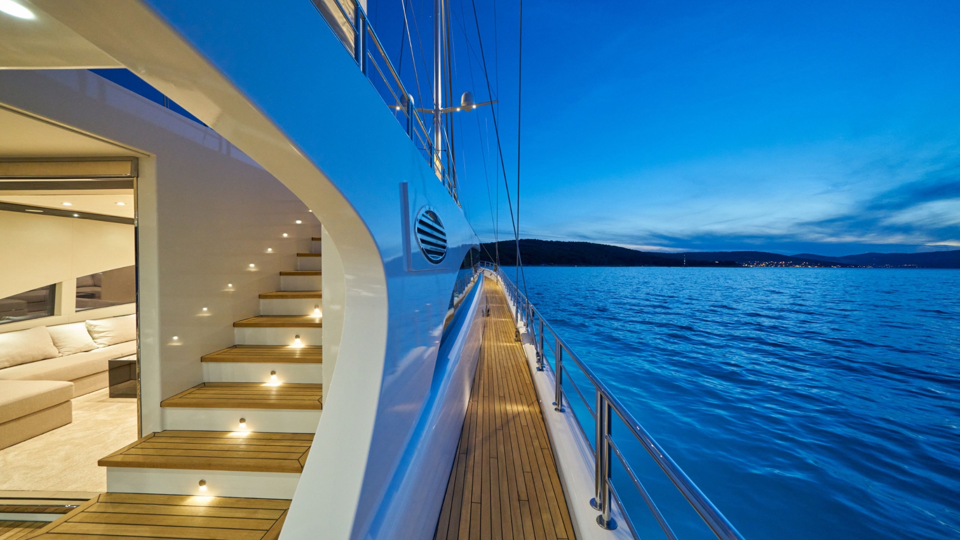 Modern yacht deck at sunset with wooden steps lit by small lights, leading inside to a lounge area. Calm blue sea and distant coastline are visible under a clear evening sky.