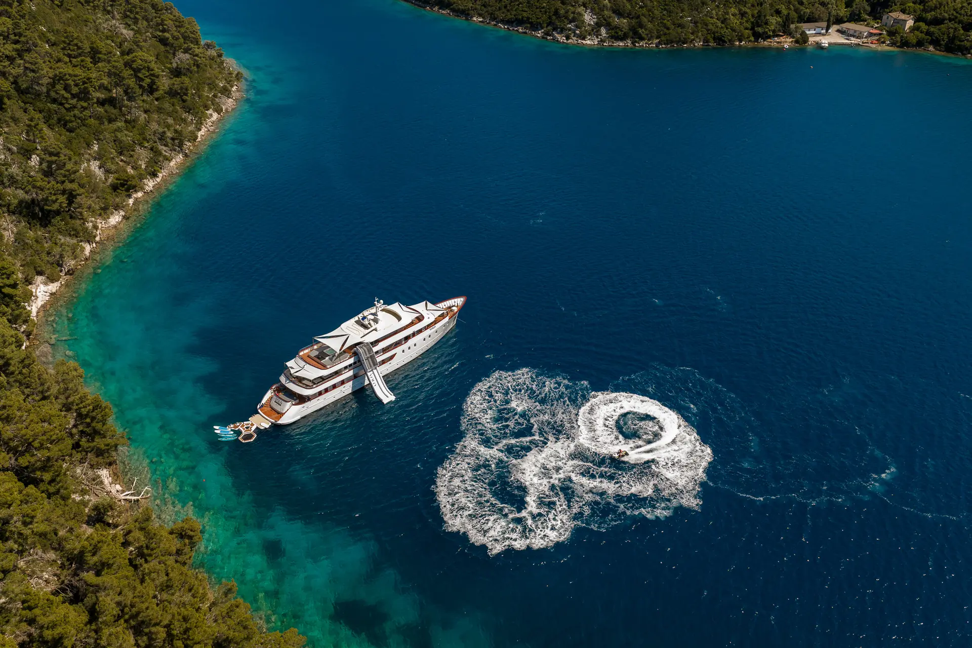 Aerial view of a large yacht anchored near a forested coastline in clear blue water, with a circular wake created by a smaller boat or jet ski nearby.
