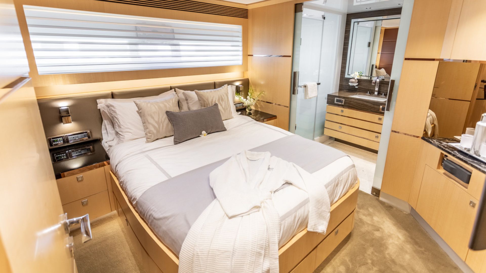 Modern yacht bedroom with light wood walls, a double bed with gray and white bedding, bathrobe on the bed, built-in storage, and a view into a sleek bathroom with a large mirror and glass shower.