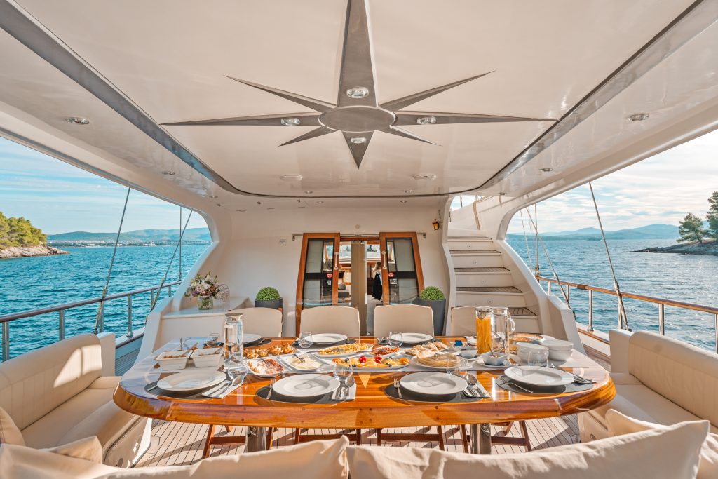 A luxurious yacht dining area set for a meal, with elegant tableware, various dishes, and drinks. The seating surrounds a polished wooden table, and the yacht overlooks blue sea and distant islands under a clear sky.