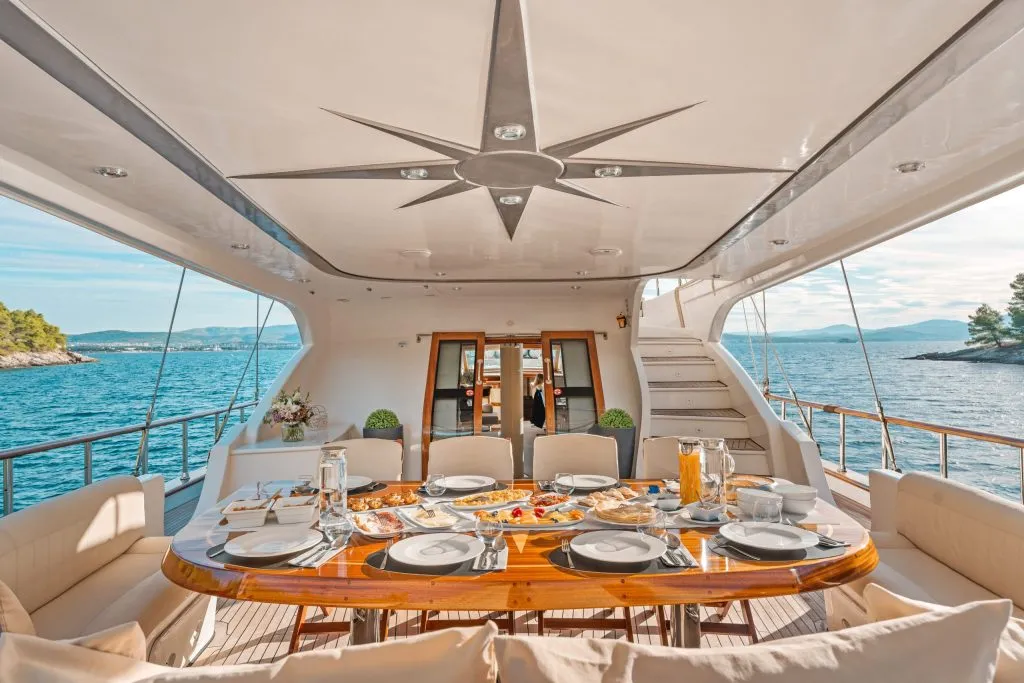A luxurious yacht dining area set for a meal, with elegant tableware, various dishes, and drinks. The seating surrounds a polished wooden table, and the yacht overlooks blue sea and distant islands under a clear sky.