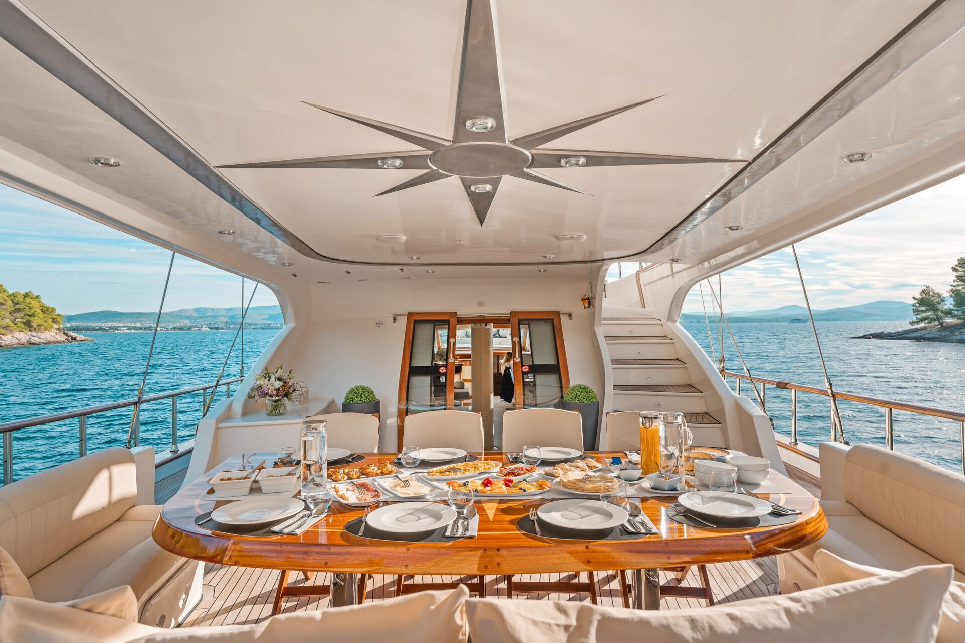 A luxurious yacht dining area set for a meal, with elegant tableware, various dishes, and drinks. The seating surrounds a polished wooden table, and the yacht overlooks blue sea and distant islands under a clear sky.