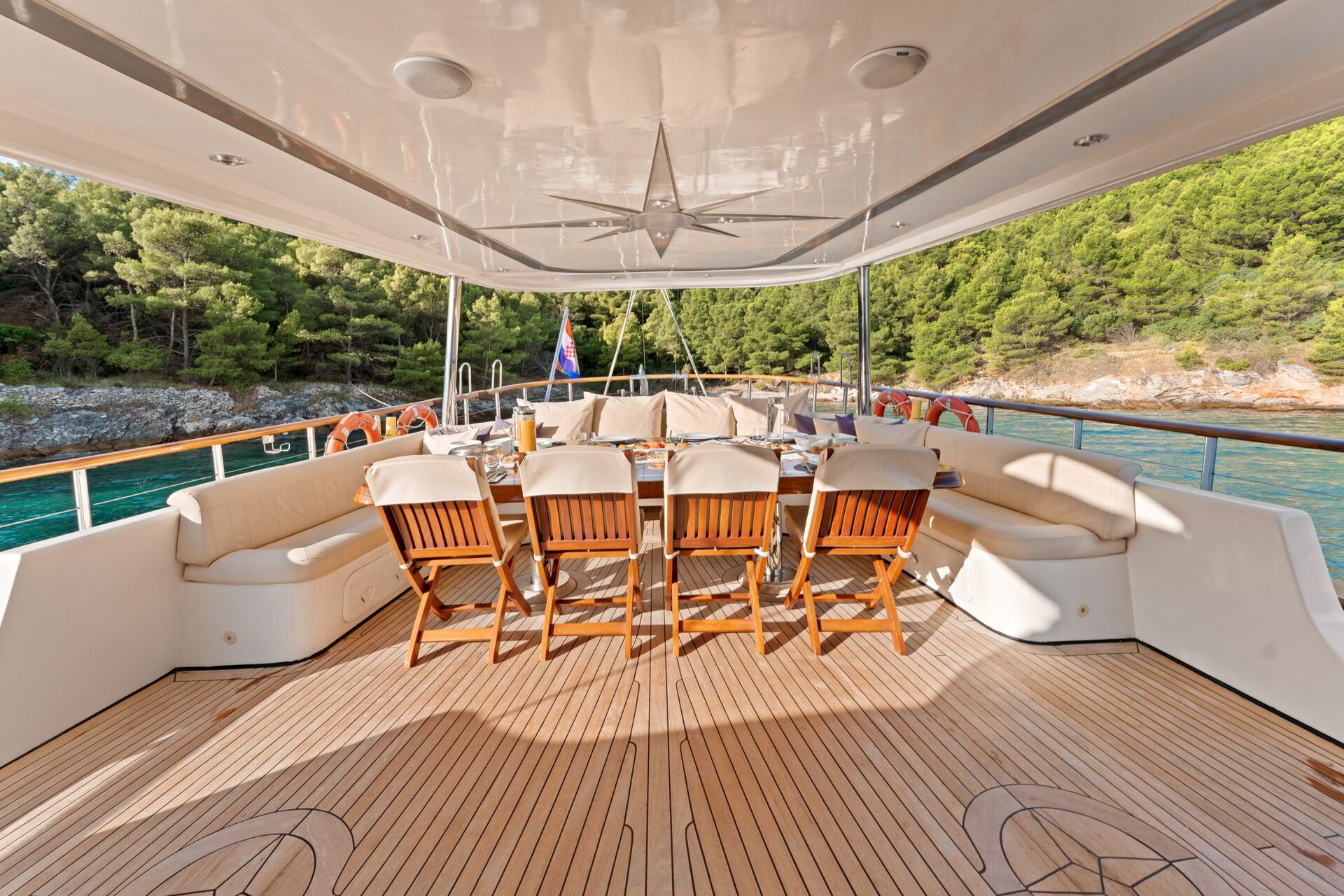 A yacht deck with a large dining table, wooden chairs, and cushioned seating. The deck overlooks clear blue water and a forested shoreline under a shaded roof. Table is set for a meal.