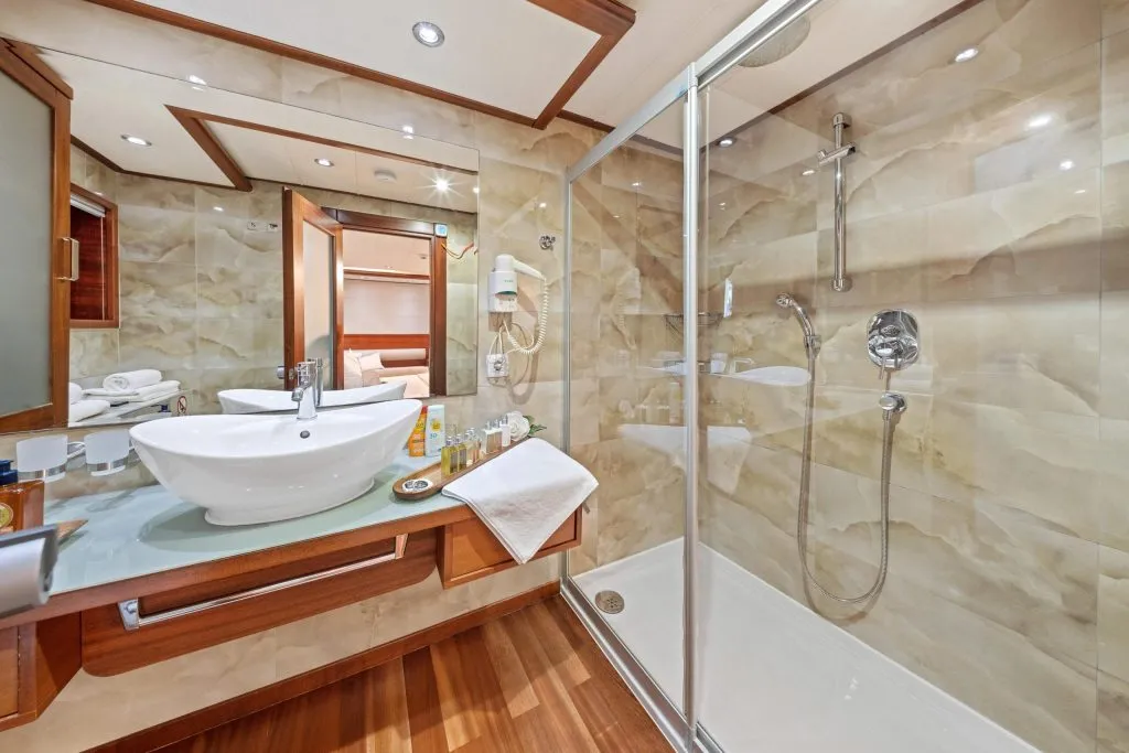 Modern bathroom with a glass-enclosed shower, wall-mounted showerhead, vessel sink on a wooden vanity, large mirror, white towels, and toiletries on the counter. Warm beige walls and wood floors create a cozy atmosphere.