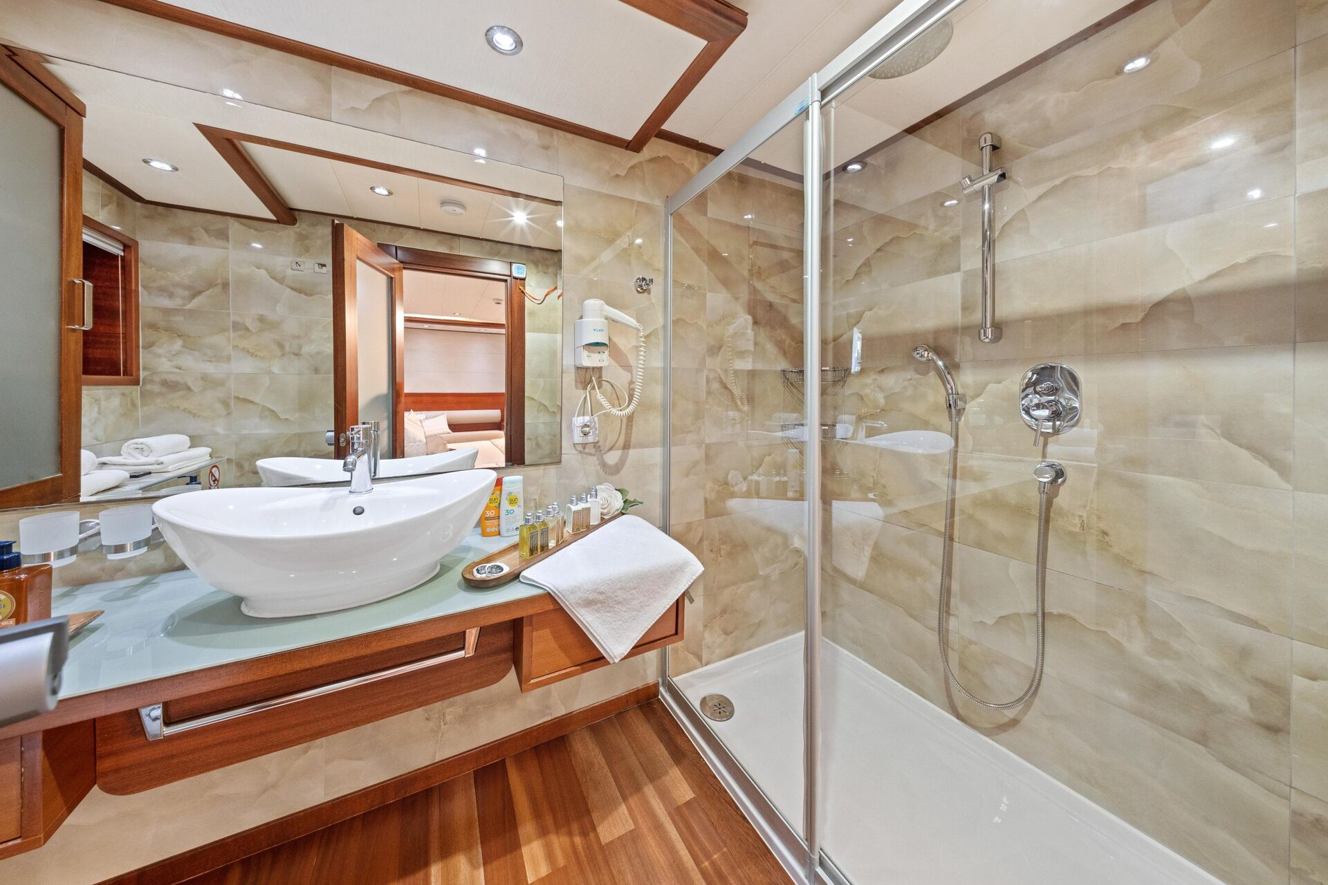 Modern bathroom with a glass-enclosed shower, wall-mounted showerhead, vessel sink on a wooden vanity, large mirror, white towels, and toiletries on the counter. Warm beige walls and wood floors create a cozy atmosphere.