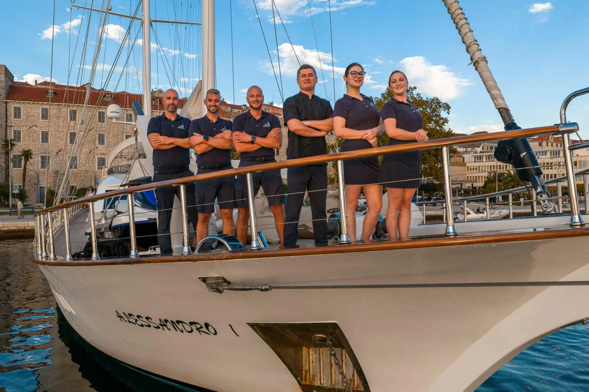 Six people stand in uniform on the deck of a white yacht, docked in a marina with buildings, trees, and a blue sky in the background. The group smiles and poses confidently for the camera.