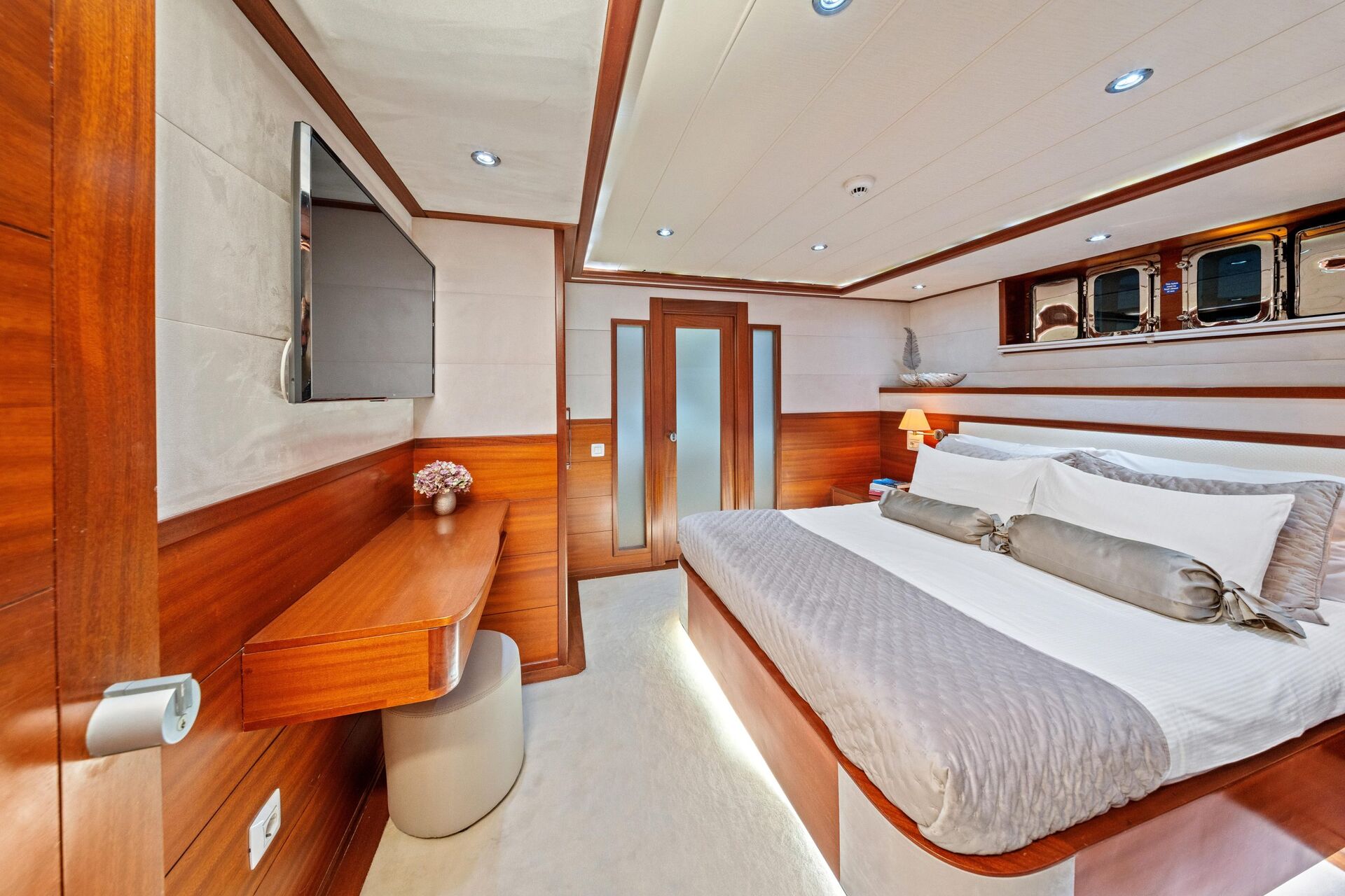 A luxurious yacht bedroom with wood-paneled walls, a large bed with gray and white bedding, a wall-mounted TV, a desk with a stool, and round windows above the headboard letting in natural light.