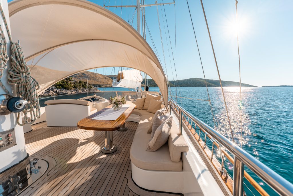Luxurious yacht deck with curved seating, a wooden table, and decorative pillows under a shade canopy, overlooking calm blue sea and distant hills on a sunny day.