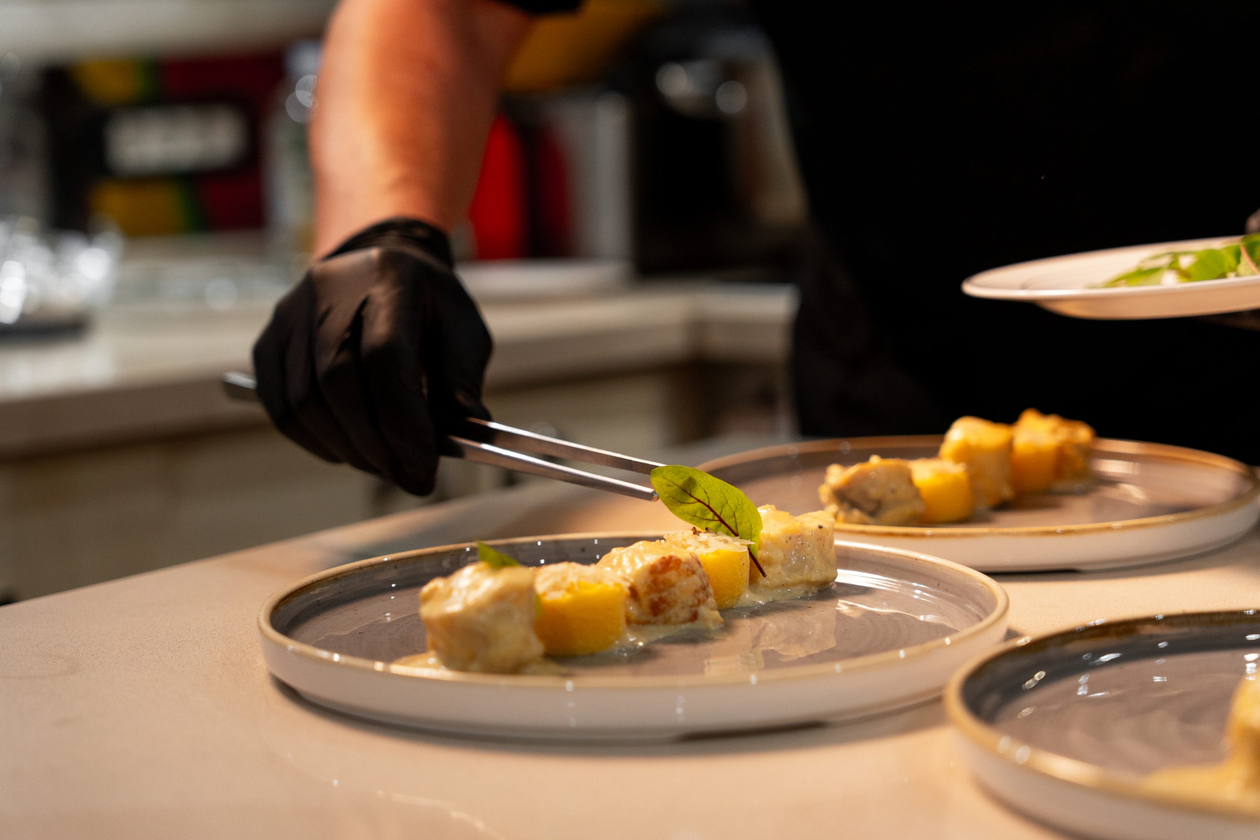 A chef wearing black gloves uses tongs to carefully place garnish on a plated dish with small portions of food in a bright kitchen setting.