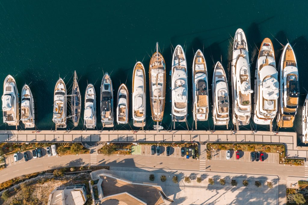 Aerial view of a marina with a row of yachts and boats docked along a pier, with a road and parked cars parallel to the waterfront. The water is deep blue and trees line the sidewalk.