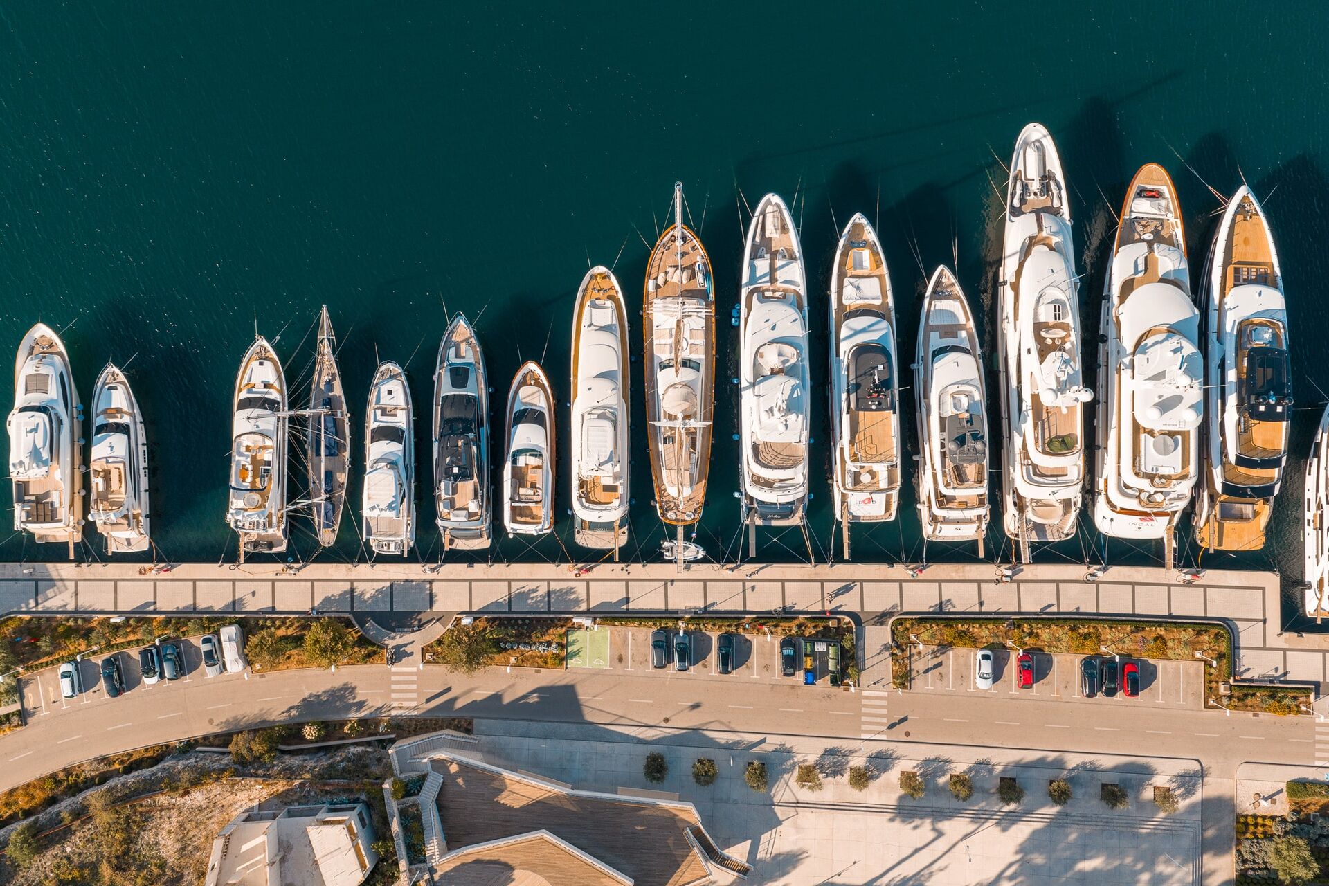 Aerial view of a marina with a row of yachts and boats docked along a pier, with a road and parked cars parallel to the waterfront. The water is deep blue and trees line the sidewalk.