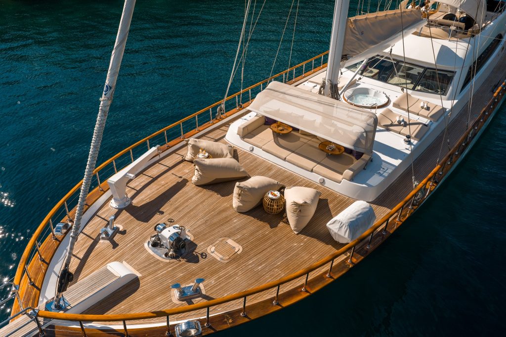 A luxury yacht deck with wooden flooring, several beige bean bag chairs, sun loungers, a jacuzzi, and small tables, floating on calm, deep blue water.
