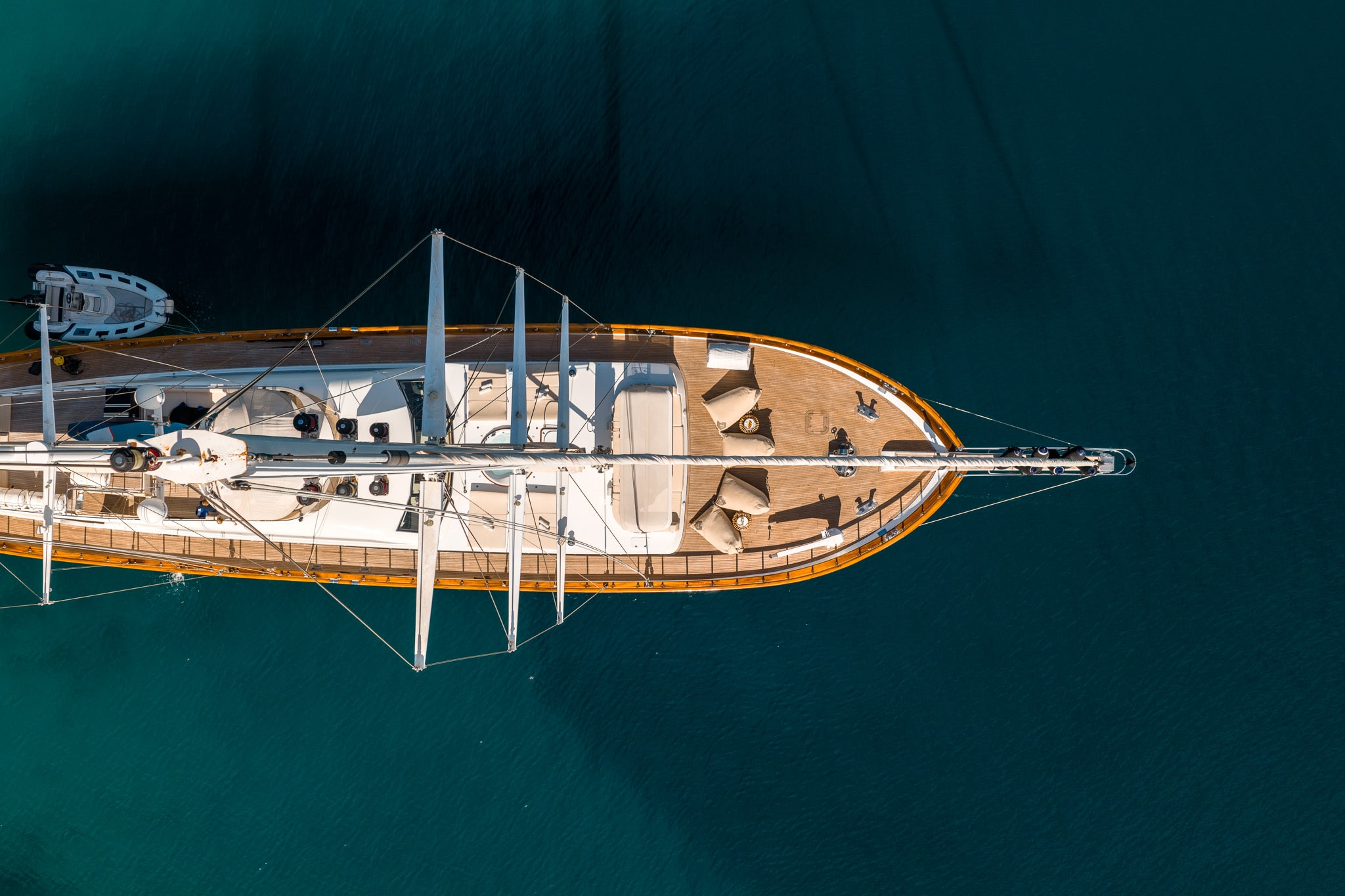 Aerial view of a large sailboat with wooden decking floating on calm, deep blue water. The boat’s lounge chairs and mast are visible, and its shadow is cast onto the water.