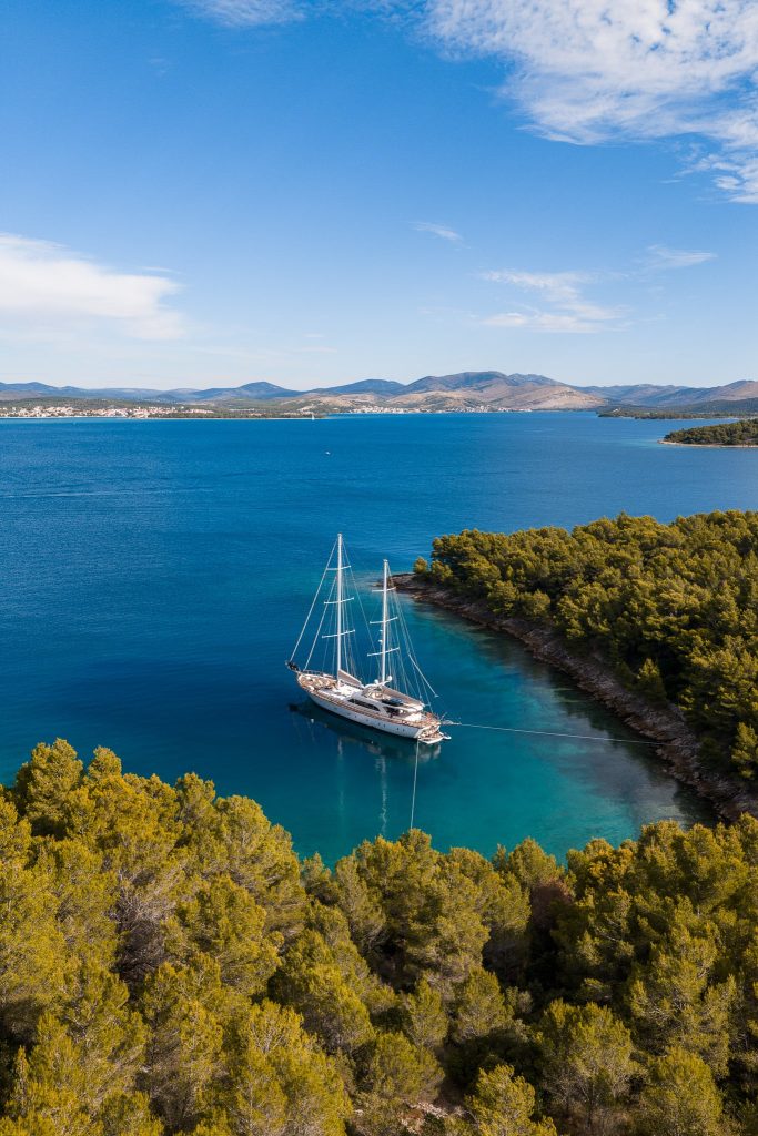 Aerial view of a sailboat anchored in a calm, blue bay surrounded by dense green trees, with distant mountains and a town visible on the horizon under a partly cloudy sky.