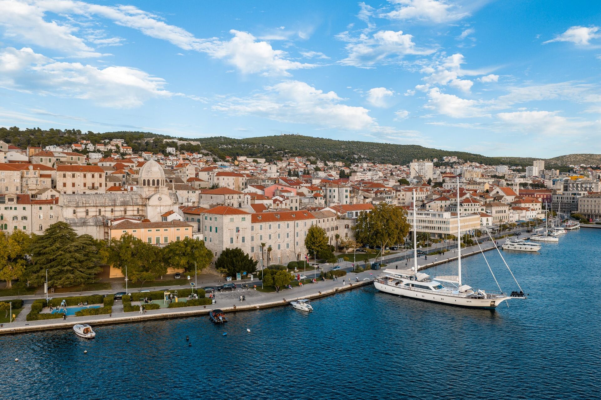 A scenic coastal town with red-roofed buildings, a marina with sailboats and yachts docked, tree-lined streets, and hills in the background under a partly cloudy blue sky.