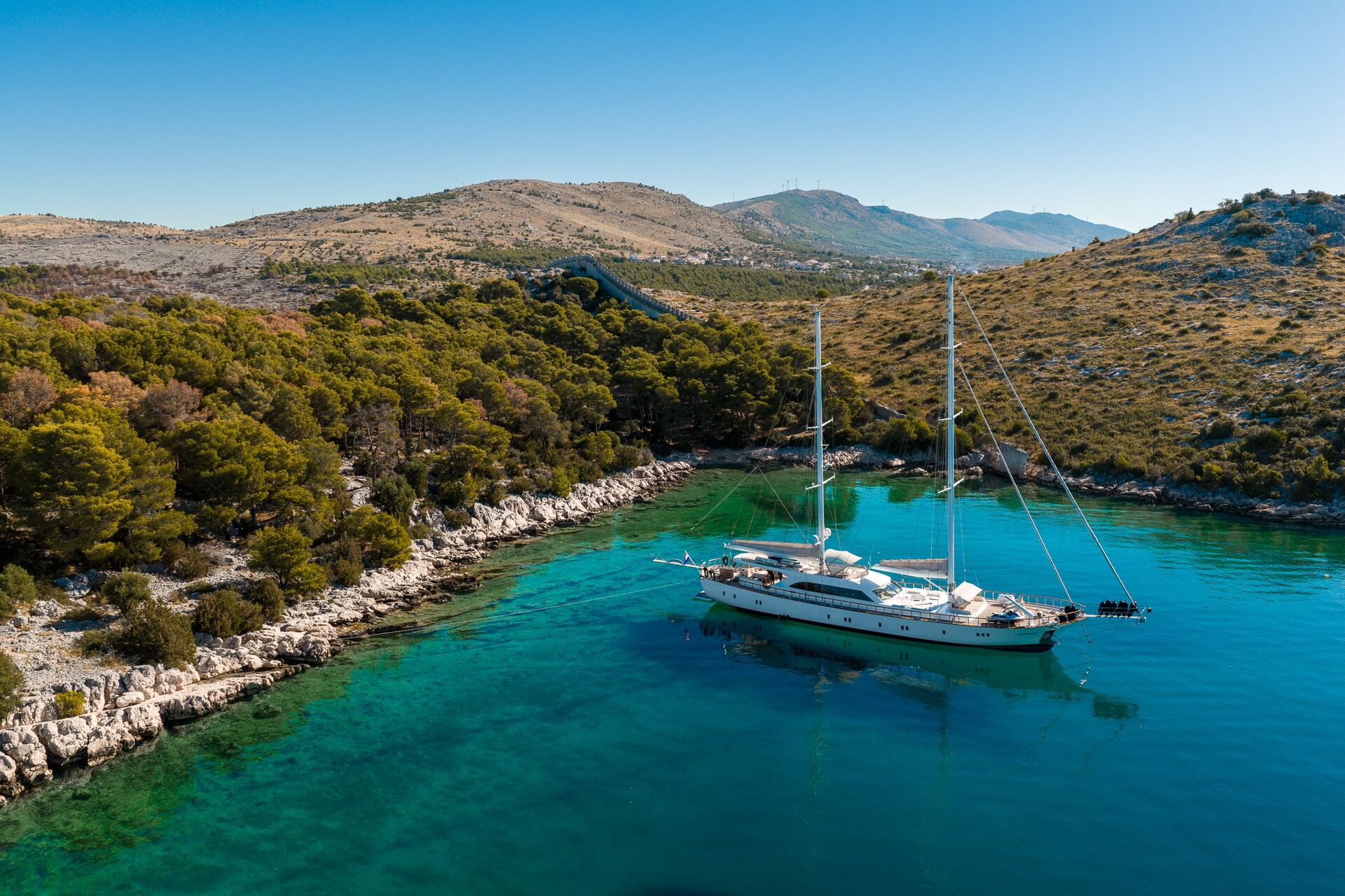 A white sailboat is anchored in a clear turquoise bay surrounded by rocky shores and lush green hills under a bright blue sky.