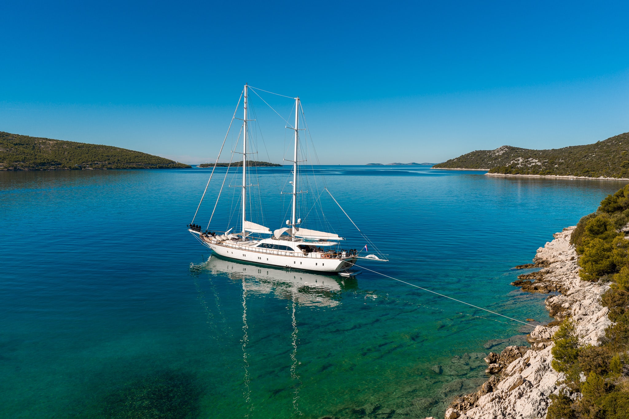 A white sailboat is anchored in clear, calm turquoise water near a rocky, green shoreline, with hilly islands in the background under a bright blue sky.