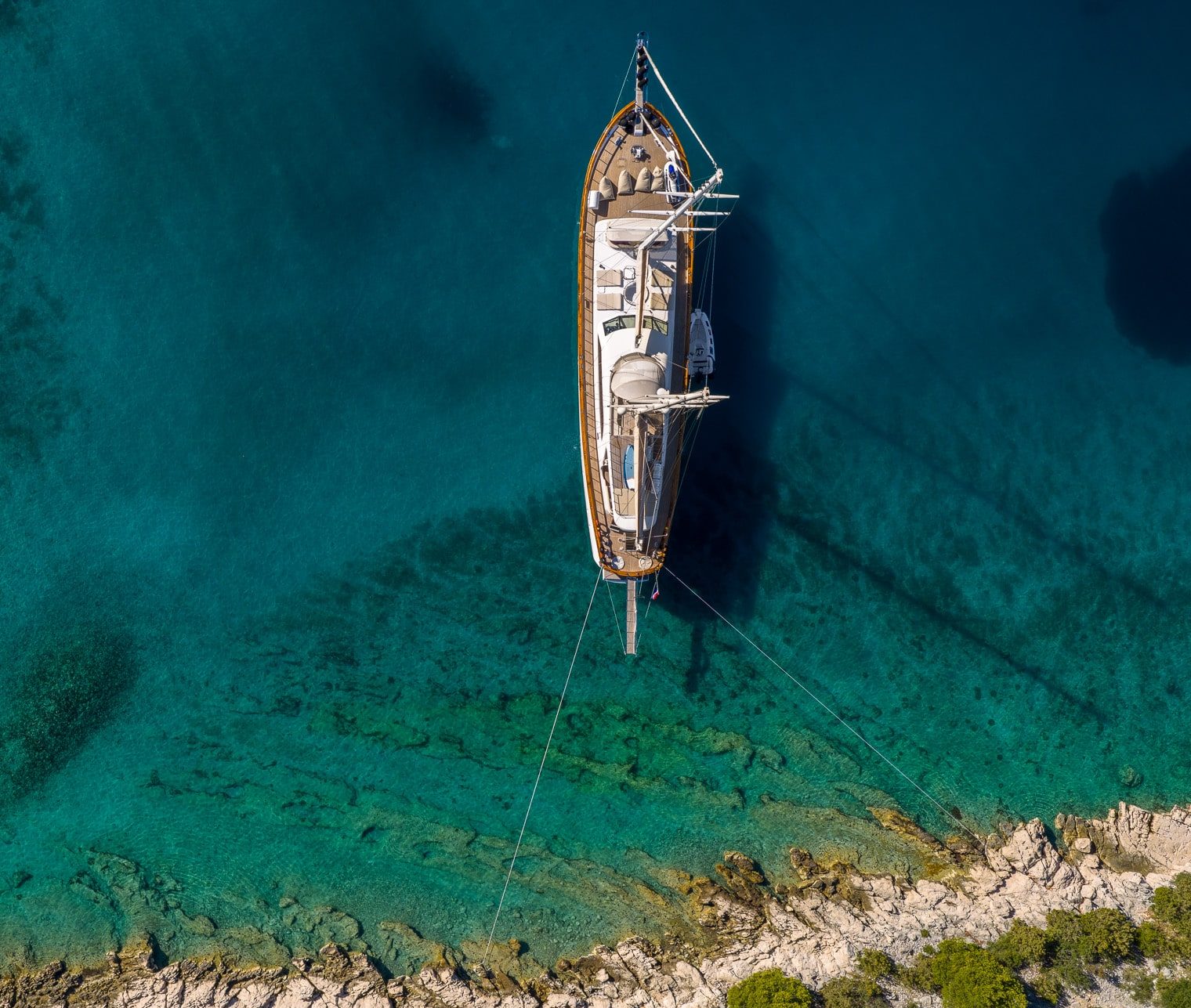 Aerial view of a sailboat anchored in clear turquoise water near a rocky coastline with green shrubs, showing the boats shadow beneath the surface.