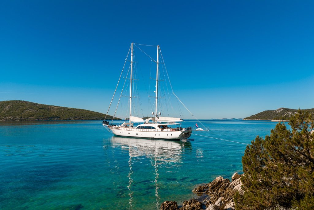 A white sailboat is anchored in clear turquoise water near a rocky shore with green shrubs, under a bright blue sky with distant hills in the background.