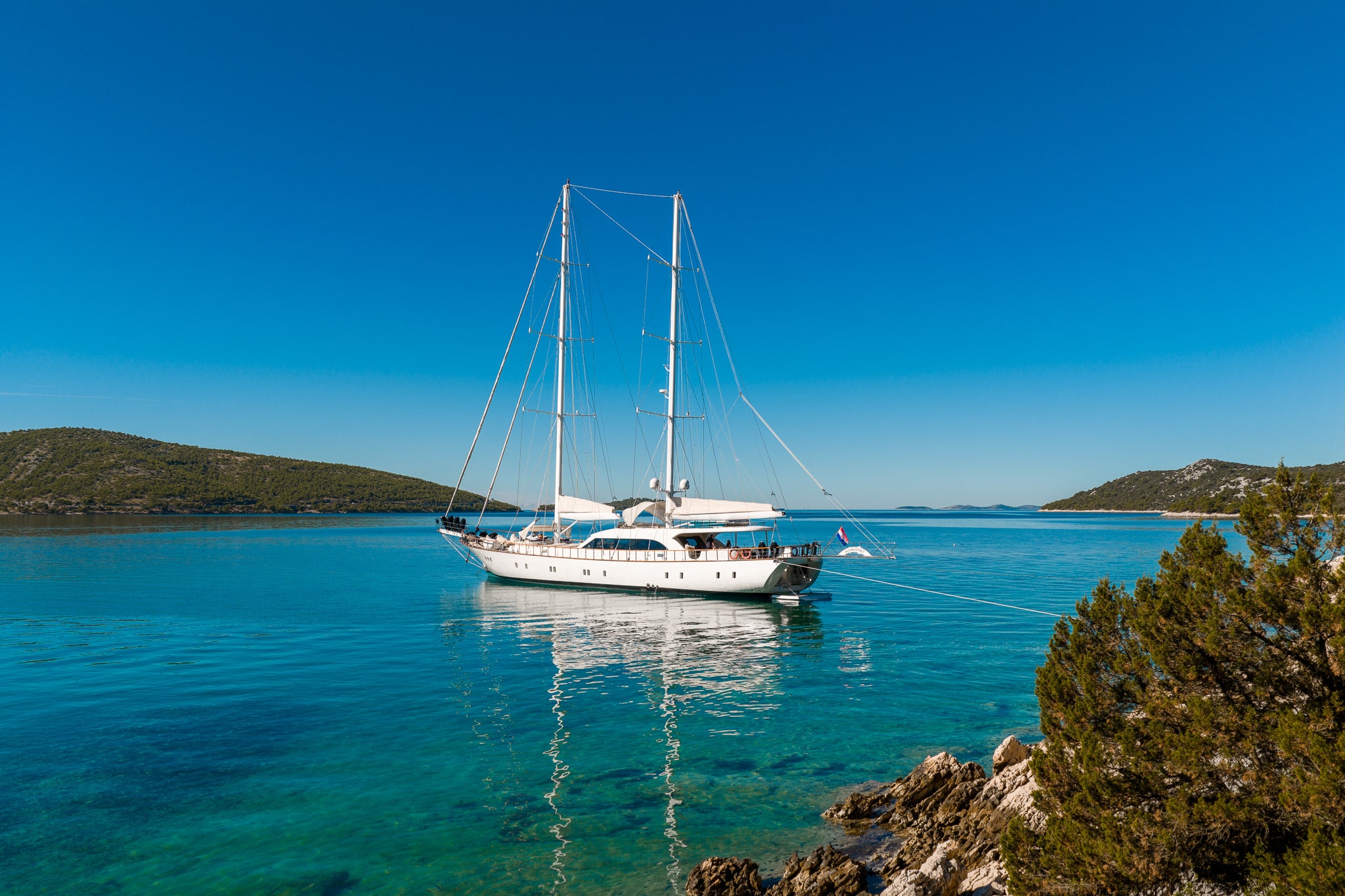 A white sailboat is anchored in clear turquoise water near a rocky shore with green shrubs, under a bright blue sky with distant hills in the background.
