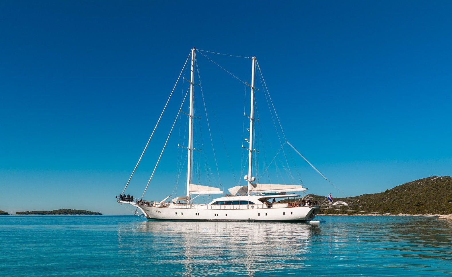 A large white sailboat is anchored on calm, clear turquoise water near a rocky shoreline, with green hills and a bright blue sky in the background.