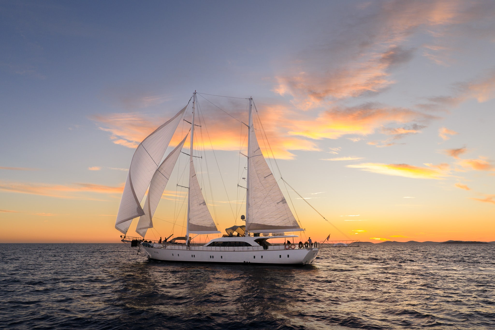 A large white sailboat with multiple sails glides across calm ocean waters at sunset, with vibrant orange and pink clouds in the sky and a distant shoreline on the horizon.
