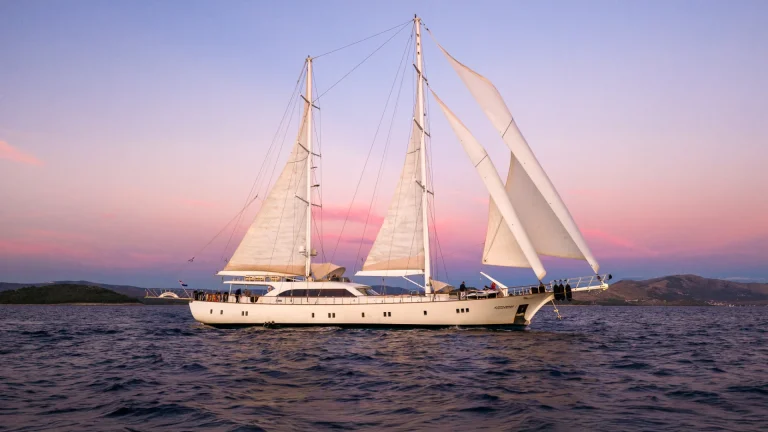 A large white sailboat with multiple sails glides on calm blue water at sunset, with a pastel pink and purple sky and distant hills in the background.