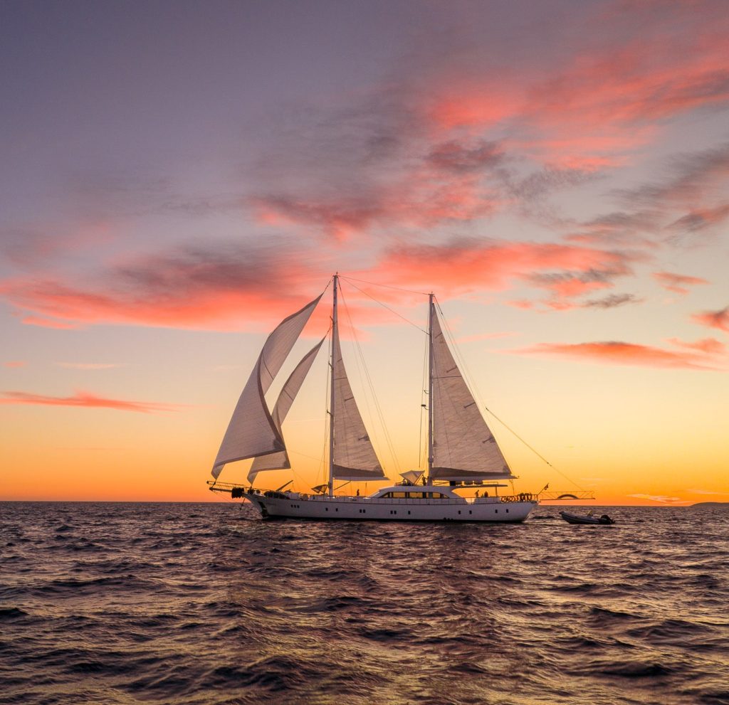 A large sailboat with white sails glides across calm ocean water at sunset, with vibrant pink and orange clouds in the sky.