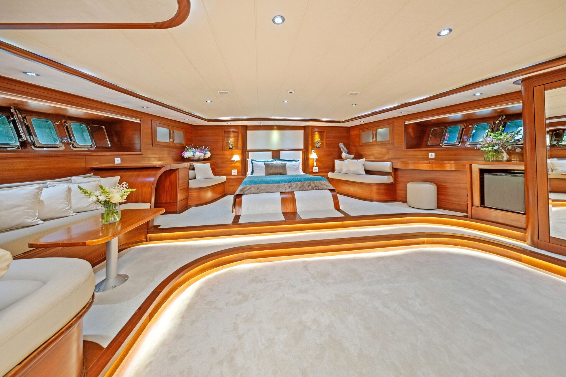 Luxurious yacht cabin with a large bed, elegant wood paneling, soft lighting, built-in seating, and flowers on tables. Multiple windows line the walls, allowing natural light to fill the spacious room.