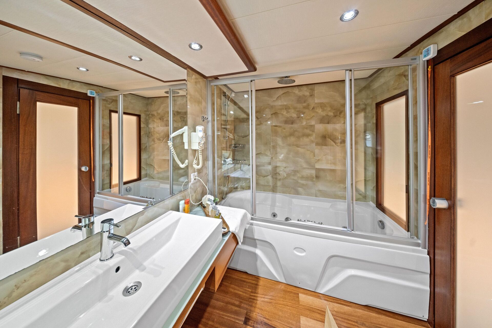 Modern bathroom with a large white sink, wooden accents, glass-enclosed bathtub and shower, wall-mounted hairdryer, and mirrors reflecting the space.