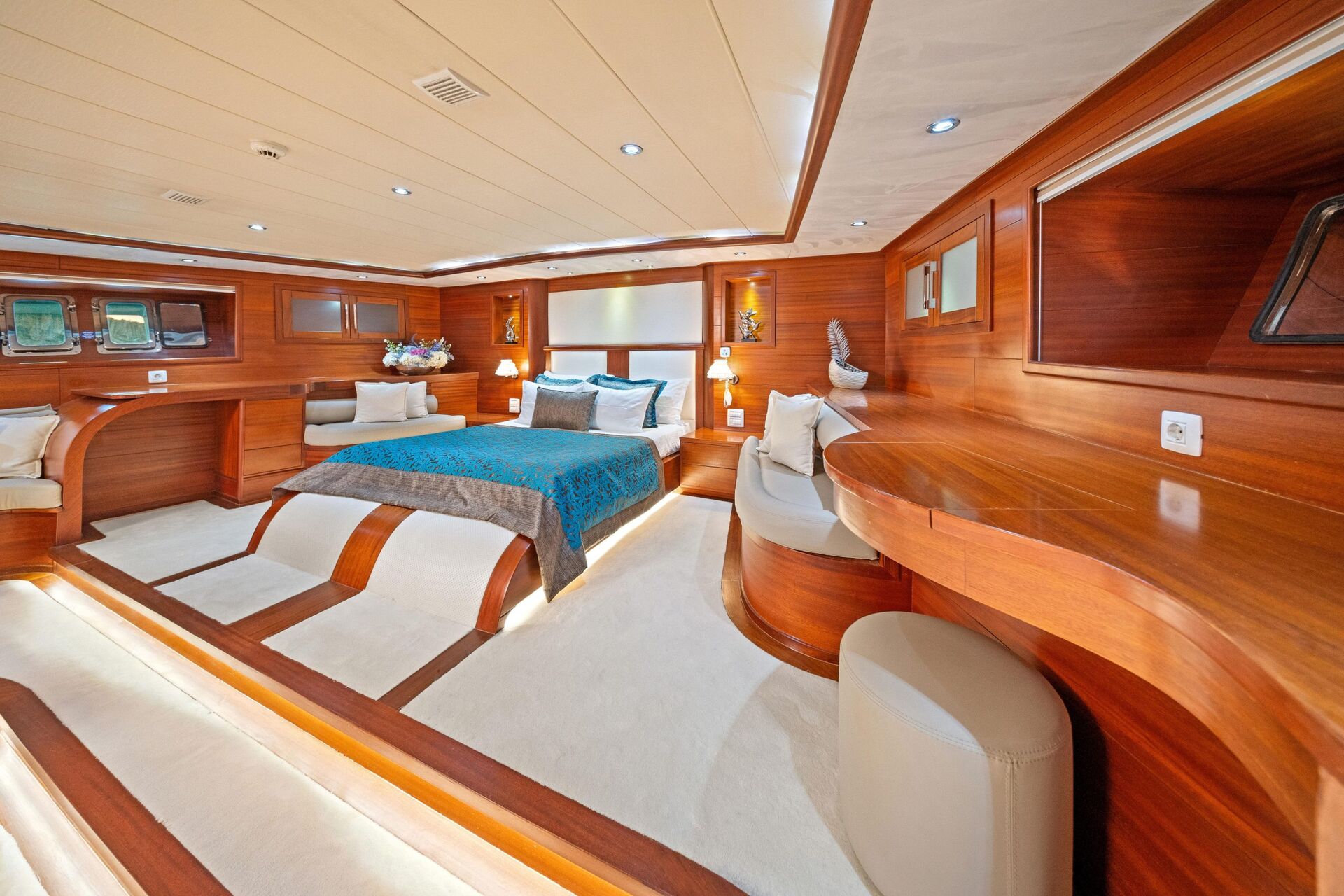 Luxurious yacht bedroom with a large bed, wooden walls and furniture, soft lighting, built-in shelves, decorative items, and plush white carpeting, creating an elegant and cozy atmosphere.