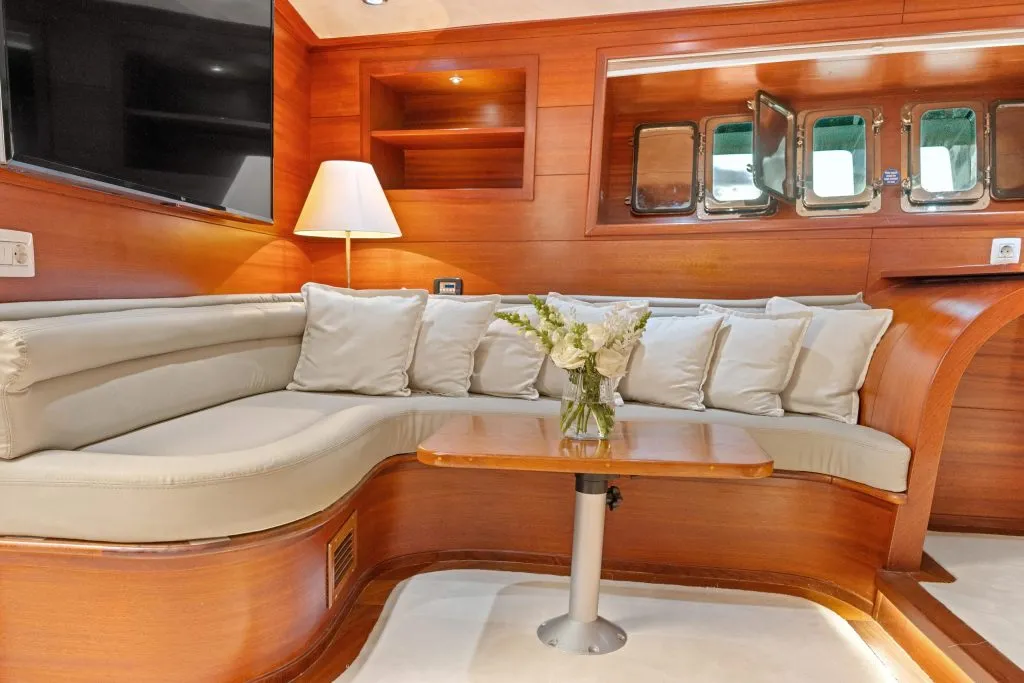 A cozy yacht interior with a curved beige sofa lined with cushions, a wooden table holding a vase of flowers, warm wood-paneled walls, a lamp, and rectangular windows reflecting greenery outside.