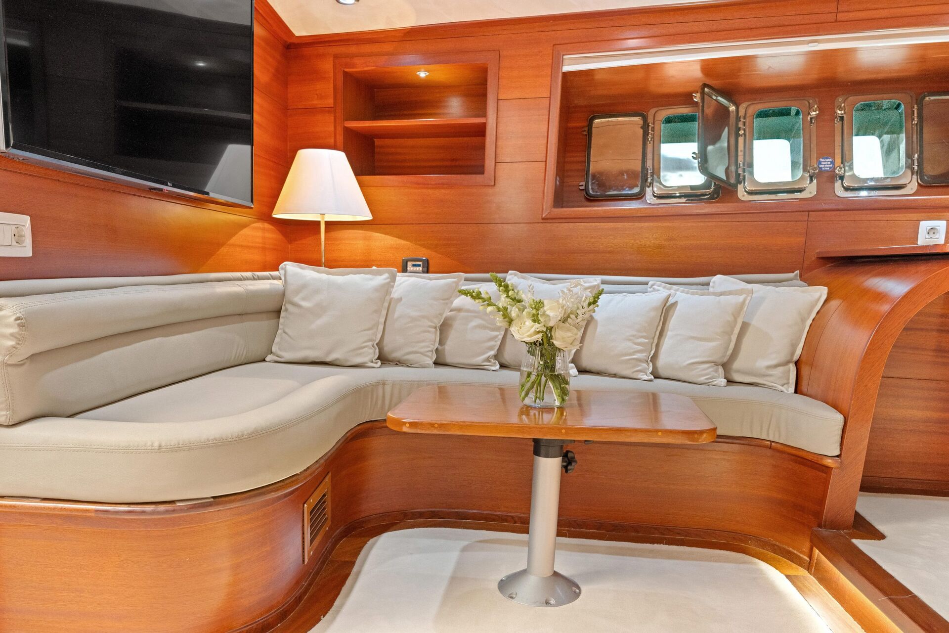 A cozy yacht interior with a curved beige sofa lined with cushions, a wooden table holding a vase of flowers, warm wood-paneled walls, a lamp, and rectangular windows reflecting greenery outside.
