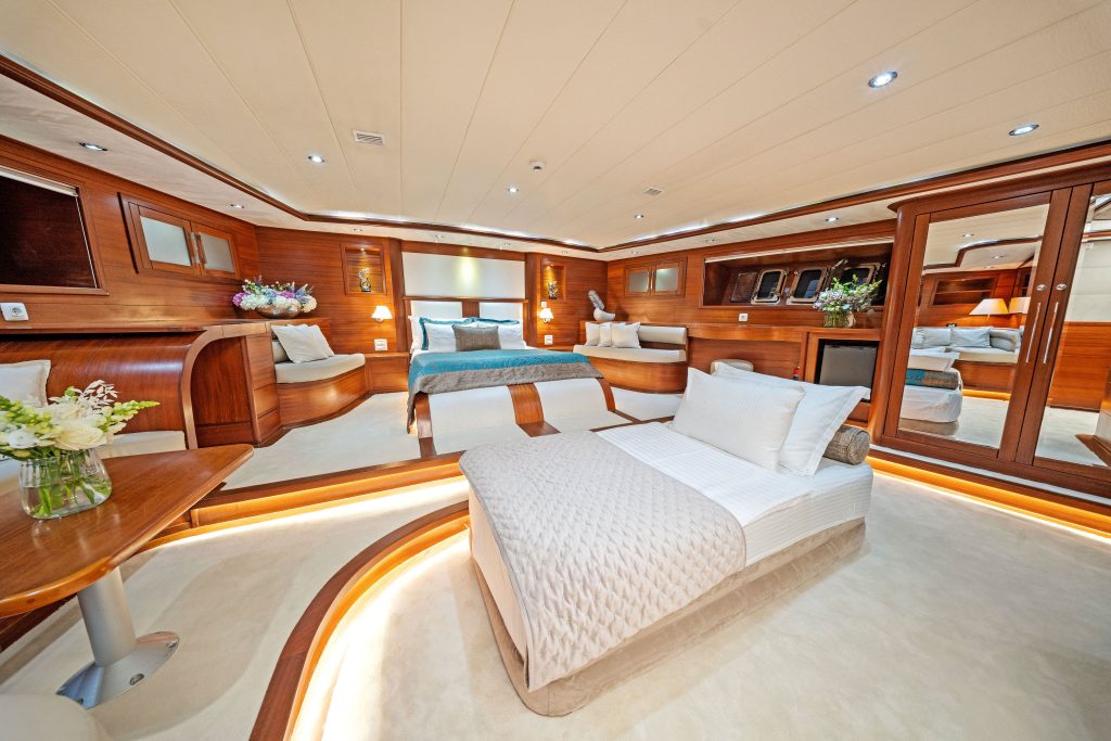 A luxurious yacht bedroom with wood paneling, a large bed, a single bed, plush white bedding, flowers on tables, ambient lighting, and mirrors reflecting the elegant, spacious interior.