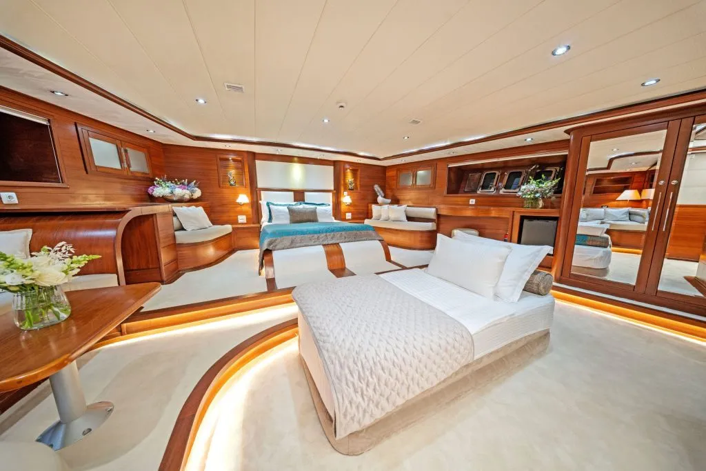 A luxurious yacht bedroom with wood paneling, a large bed, a single bed, plush white bedding, flowers on tables, ambient lighting, and mirrors reflecting the elegant, spacious interior.