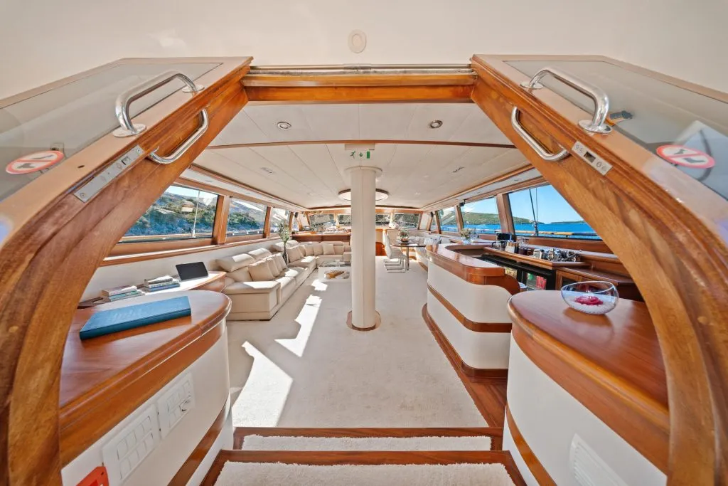 View from an open doorway into a luxurious yacht interior featuring cream-colored seating, a bar area, large windows with scenic ocean views, and polished wooden trim throughout.