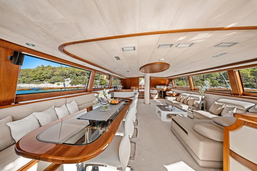 Luxurious yacht interior with plush cream sofas, elegant wood accents, a long dining table, and large windows offering views of the sea and trees outside. Vases with flowers and fruit decorate the space.