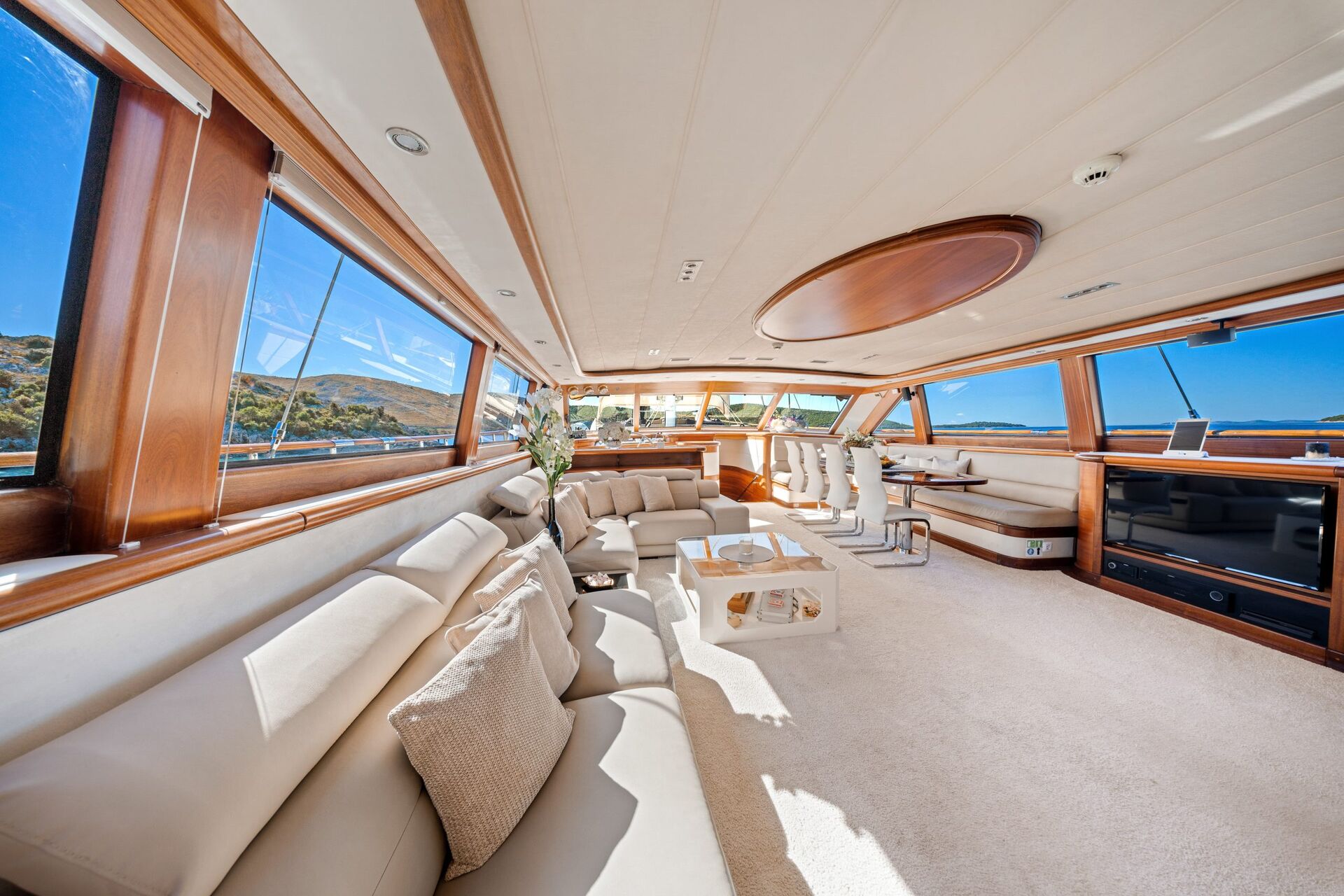 Luxurious yacht interior with large beige sofas, cushions, a coffee table, and wide windows offering scenic water and landscape views under bright blue skies. Natural light fills the spacious, elegant setting.